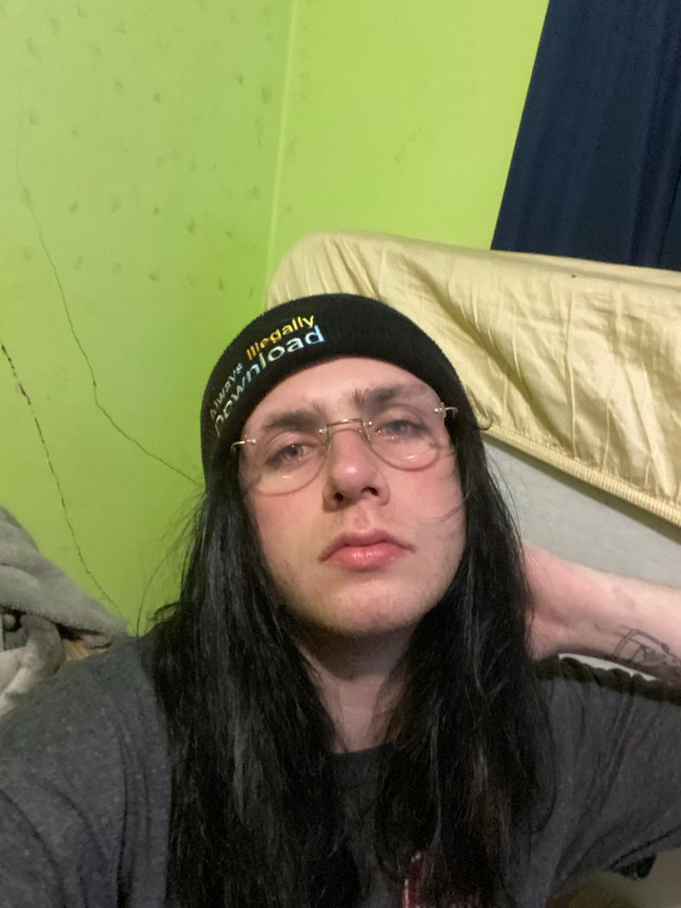 A stoner rabbit posing in their room. They are wearing rimless glasses, a grey t-shirt and a beanie that says “always illegally download” 