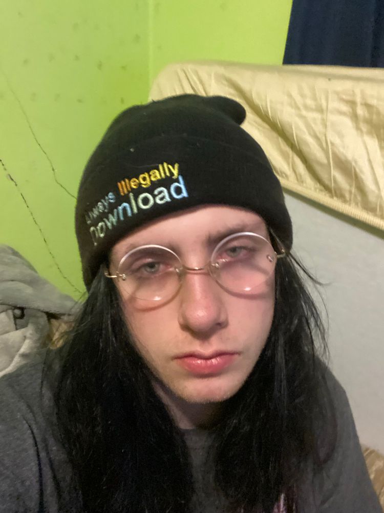 A stoner rabbit posing in their room. They are wearing rimless glasses, a grey t-shirt and a beanie that says “always illegally download” 