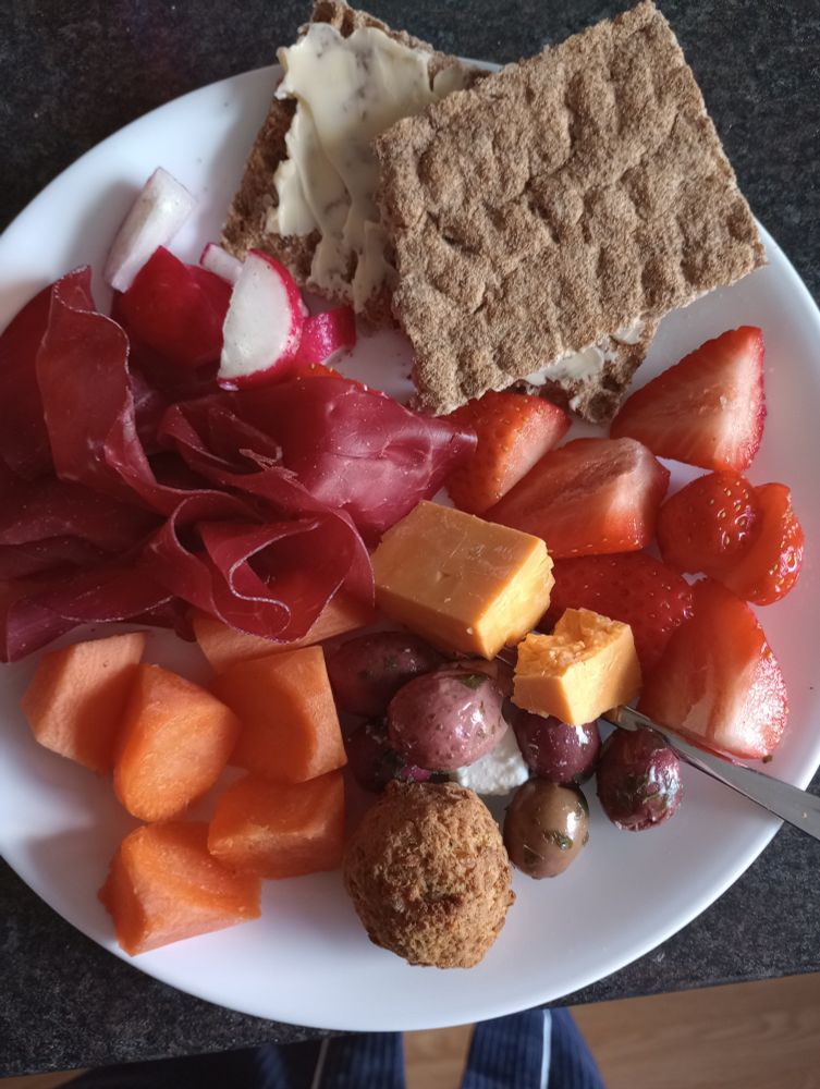 A white plate with various foods arranged on it. Buttered rye crispbread, sliced strawberries, marinated black olives with feta, a couple chunks of red Leicester cheese, a falafel, chunks of carrot, bresaola, and sliced radishes.