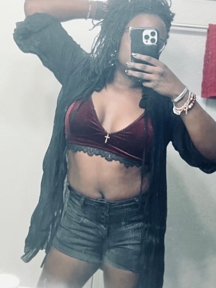 Sheron in bathroom mirror wearing black denim shorts, a velvet red bra let and a see-through blouse.