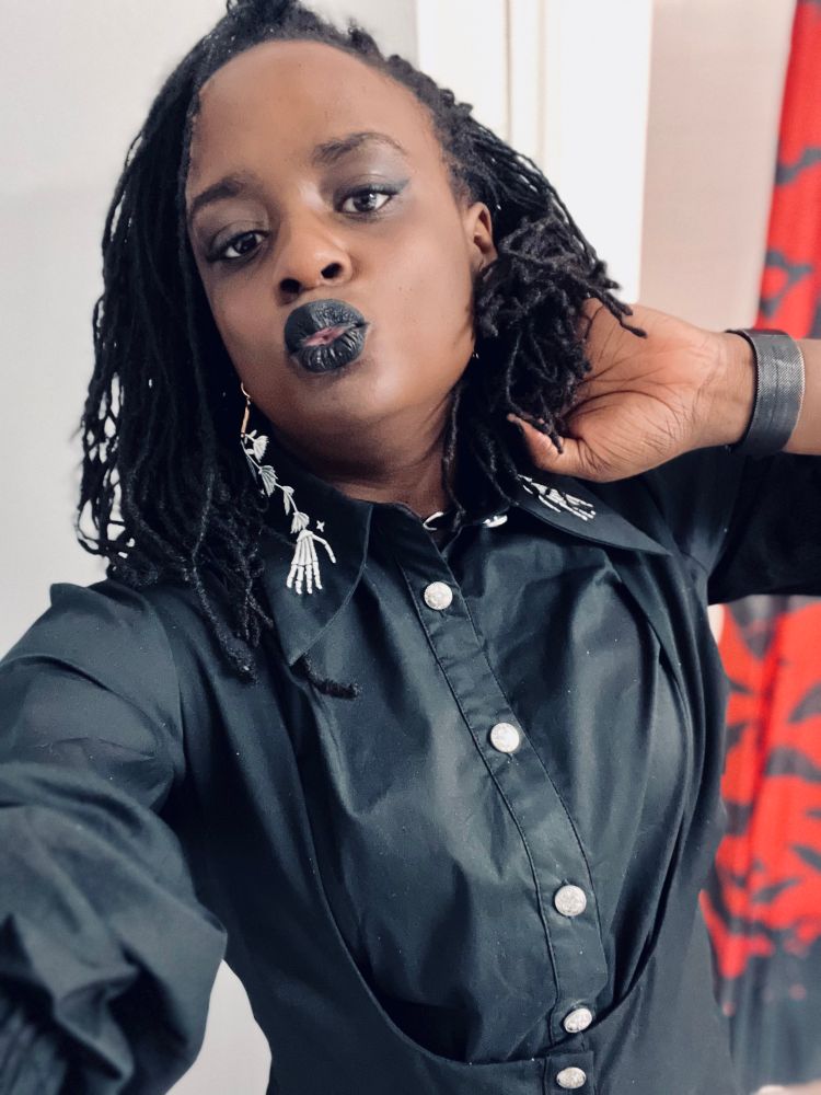 Sheron kissy facing toward the camera in her bathroom wearing a black jumpsuit and a black blouse beneath it 