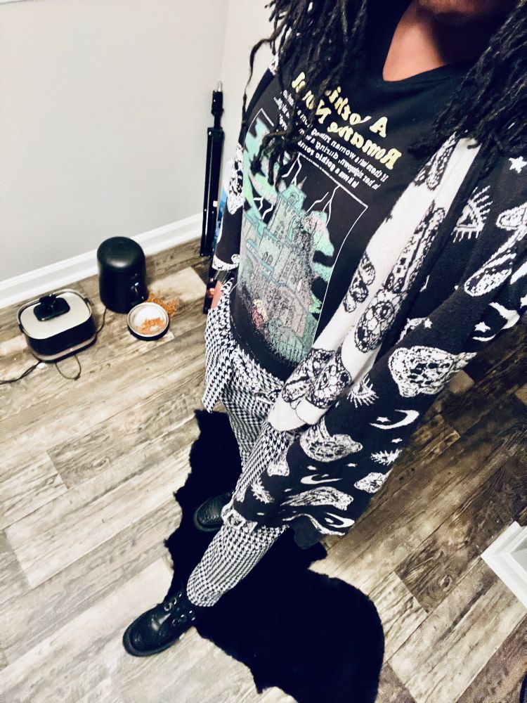 Sheron posing on top of black bat rug wearing black t.u.k.s, white and black plaid pants with a graphic tee and a black and white cardigan with magical symbols