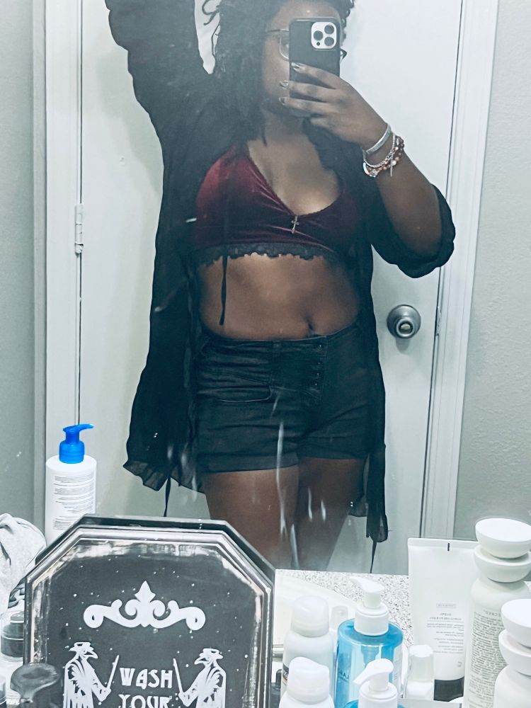 Sheron in bathroom mirror wearing black denim shorts, a velvet red bra let and a see-through blouse.
