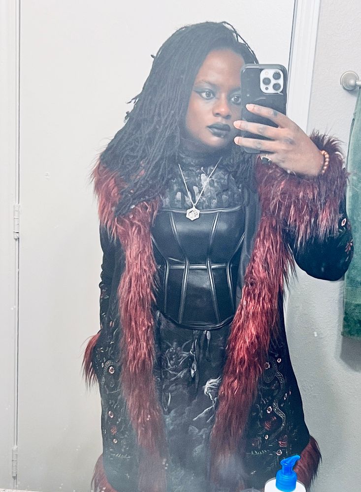 Sheron in bathroom mirror wearing black long dress with skeleton designs, and a black corset over it. Over that she is wearing a black coat with red fake fur trimming 