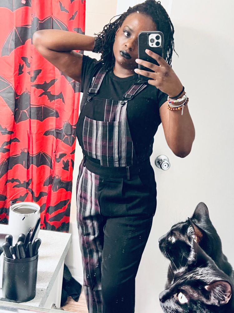 Sheron posing in bathroom in purple plaid and black overalls. Her black cats head appears mirrored in the lower right corner. 