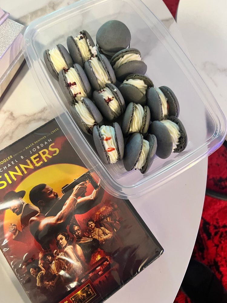 DVD of the movie sinners next to a container of strawberry flavored macarons with gray cookies and white cream middles with red blood drops in the centers. These are on top a white marble coffee table 