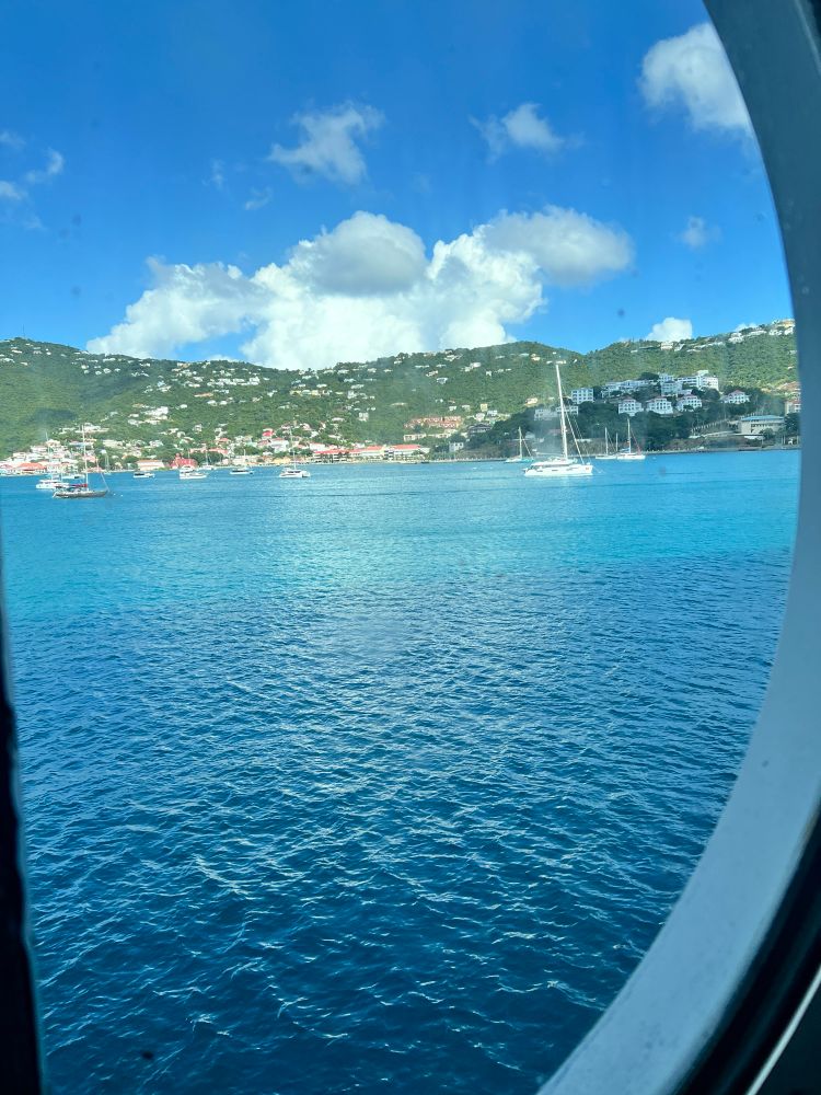 Picture of see-through round cruise ship window showing blue sea and boats of various sizes as well as the land of saint Thomas. The sky is bright blue with large, thick clouds. 