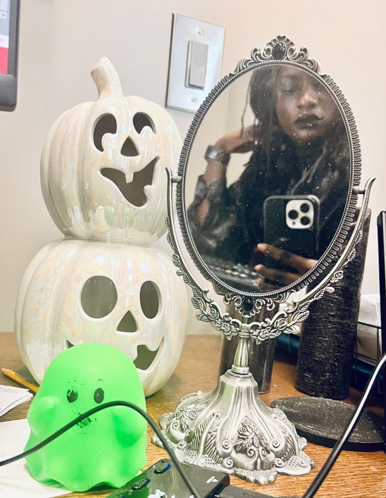 Sheron taking mirror selfie with small vintage vanity mirror on desk with a black faux candle behind it, a green ghost nee-do, and a two-stack white Jack o lantern decoration behind that. 