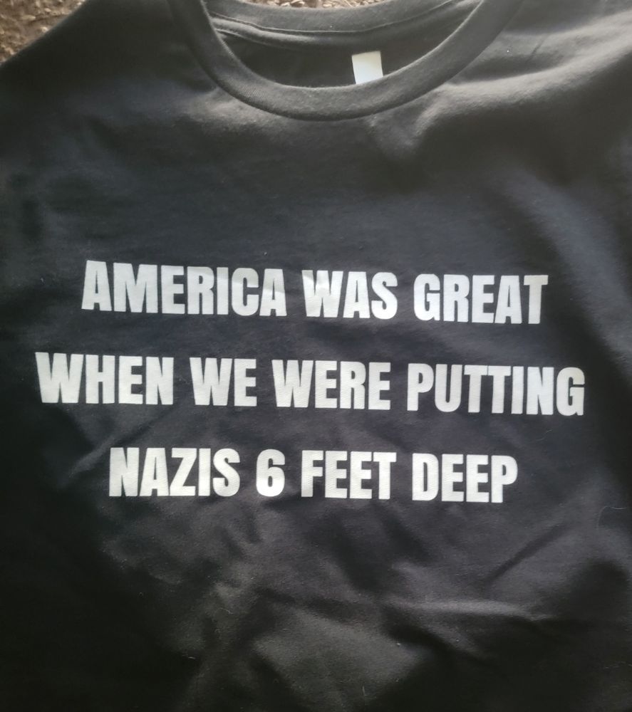 Black shirt with white text stating "America was Great when we were putting Nazis 6 feet deep" 