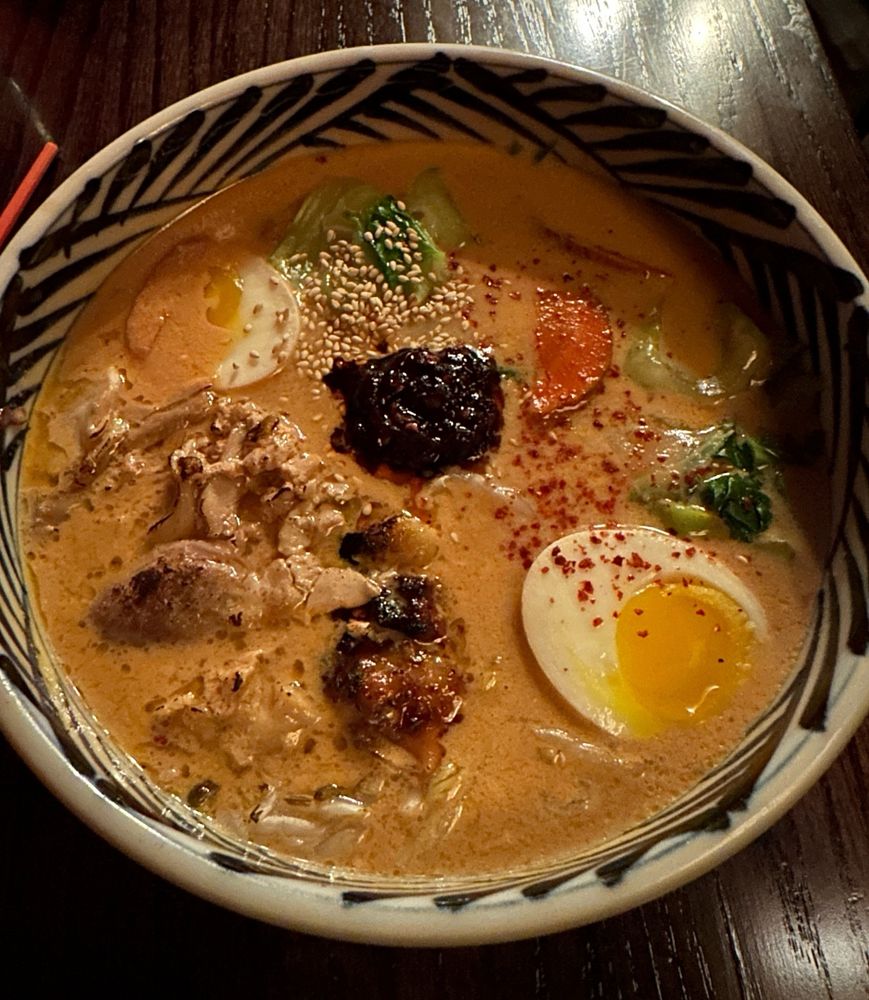 Bowl of creamy ramen with noodles, chicken, greens, and half of an egg on a table. 
