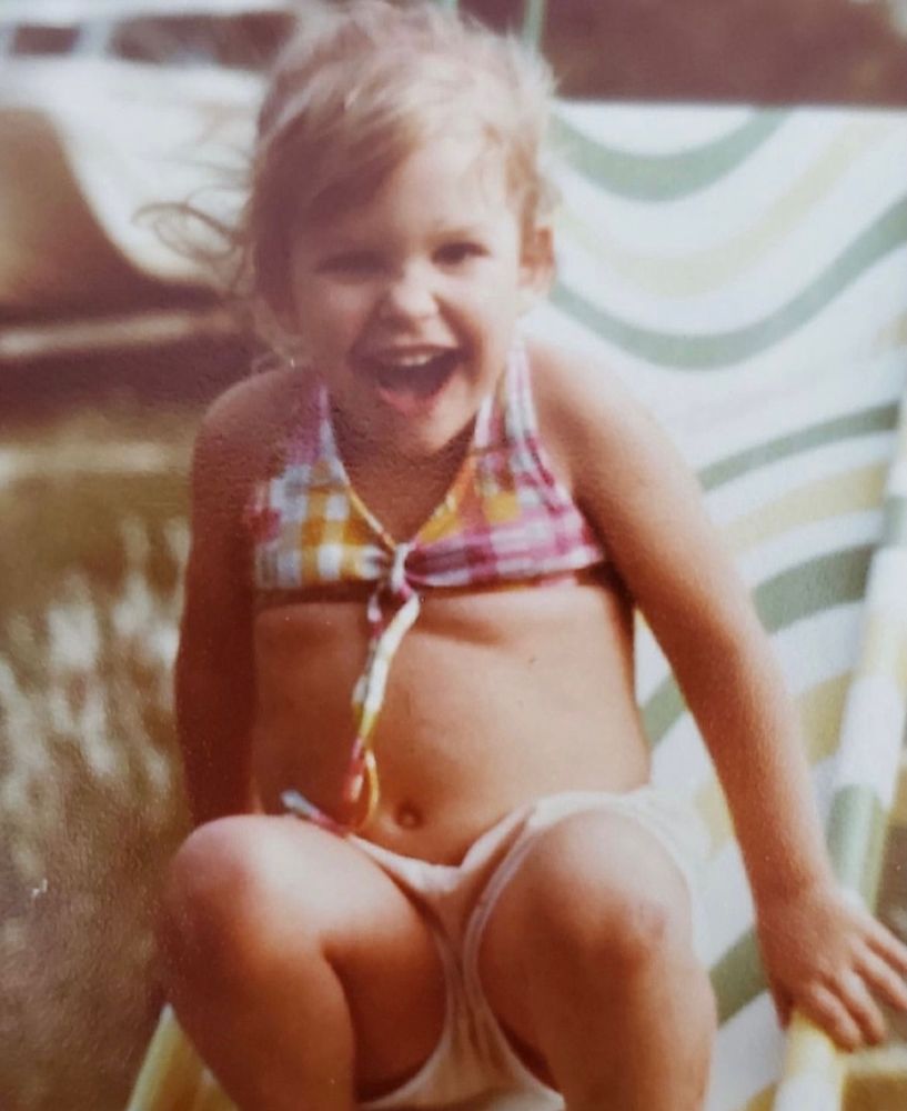 ya girl at about age 2-3 with blonde bangs, pastel swim suit top and shorts, screaming laughing going down a slide 