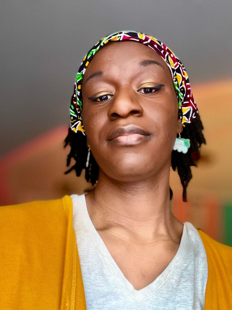 Selfie of Maya from a lower angle. She is wearing makeup with black eyeliner, gold eyeshadow, bulbasaur earrings and a colorful bandana. She is in front of a blurred orange tinted led background.