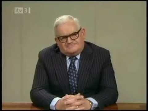 Ronnie Barker, a comedian renowned for puns and wordplay, sitting behind a desk and dressed in a suit and tie in the style of a TV news presenter