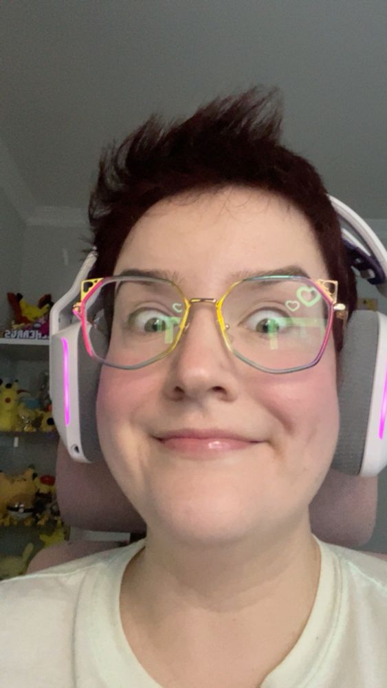 A selfie of Stef. The camera is close up and she’s smiling softly with her eyes wide open in a silly way. She’s wearing rainbow geometrical glasses, a white headset, and a light blue t shirt.