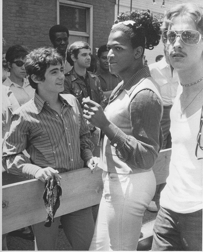 Marsha P Johnson in a skirt, button down dress shirt and best standing beside a wooden rail with several gay men.