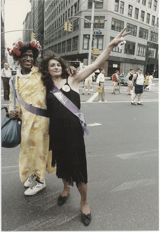Marsha P Johnson and Silvie Rivera standing together in the street. Marsha is wearing a yellow dress, and Silvie a black dress. Silvie is holding her hand up in a Peace symbol.