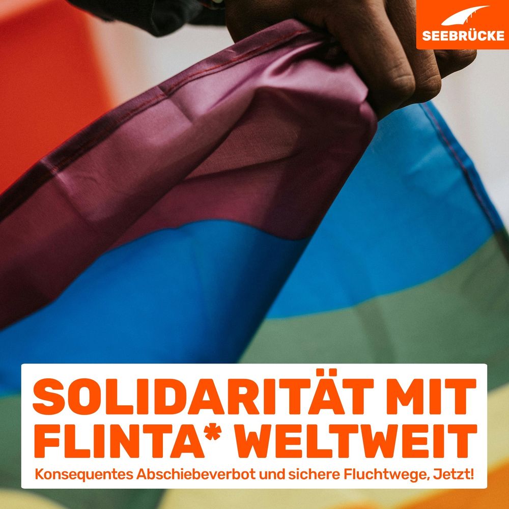 Eine Regenbogenfahne wird in einer Hand gehalten. In einem weißen Kasten steht "Solidarität mit FLINTA* weltweit. Konsequentes Abschiebeverbot und sichere Fluchtwege. Jetzt!". In der rechten oberen Ecke ist das Seebrücke-Logo in weiß auf orange.
