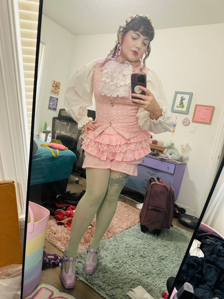 The OP standing in front of her bedroom mirror to show off a “rococo” inspired party costume. It includes a handmade cravat, pink vest, pink frilly shorts, poofy white sleeves, pale green tights, and lavender platform heels.