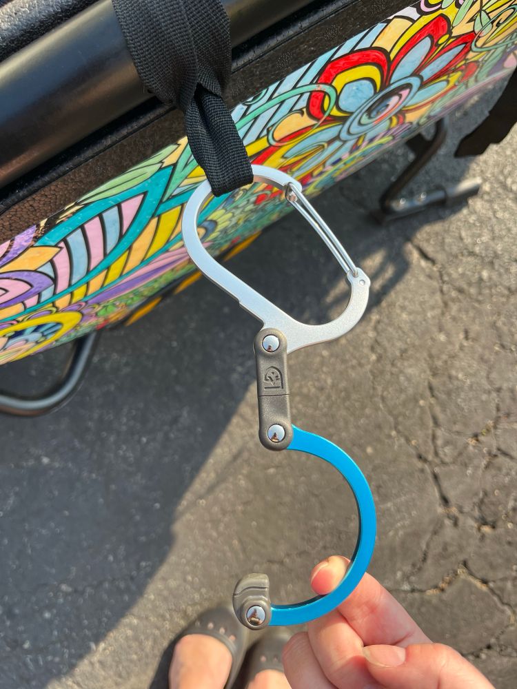 A close-up of a blue medium-sized Heroclip attached by a black nylon webbing straps to the frame of an Urban Arrow bucket.

The clip is a palm-sized carabiner clip with another additional hook attached to it with a little rubber foot at the end.