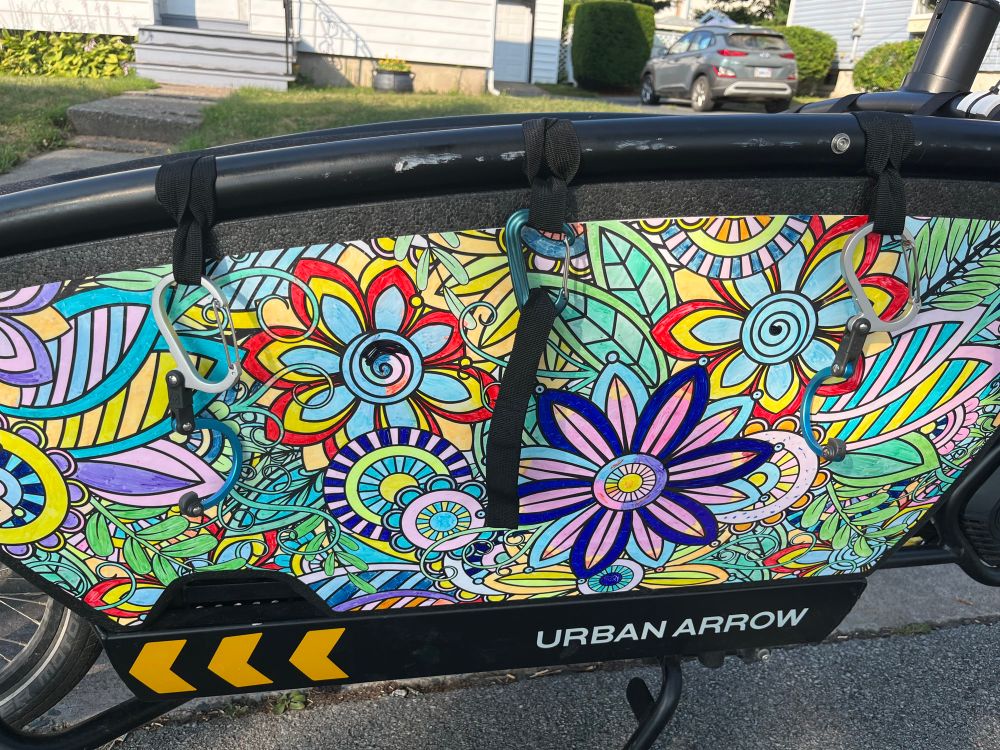 An Urban Arrow cargo bike is parked at the side of a residential street. The bucket has a colourful flower sticker on it. Short black nylon straps circle the side top tube of the box frame in three places: front, middle, and back. The front and back straps have Heroclip carabiners attached, with the hook extended to hold the tire of a child’s bike (not pictured).  A simple carabiner hangs from the middle strap, with a fourth strap attached to wrap around the middle of the bike as a safety.