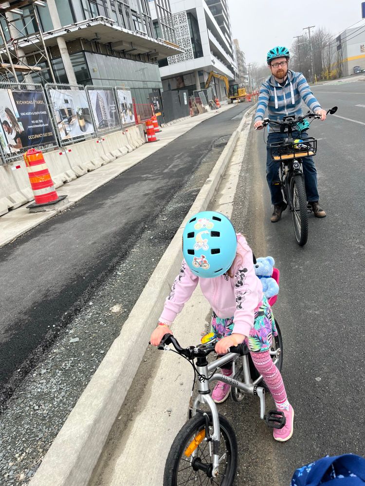 A raised asphalt bike lane runs along the left side of the photo. In the centre foreground a 5 yo girl wearing a pink hoodie is astride a silver bike, in the road next to the bike lane. She has pink leggings and purple sneakers, and a blue teddy bear sits
 in a doll seat on the back of her bike. She is wearing a teal helmet and looking down. Behind the girl her father is astride his own bike, looking grim. In the far background you can just make out some signage closing the bike lane and sidewalk.