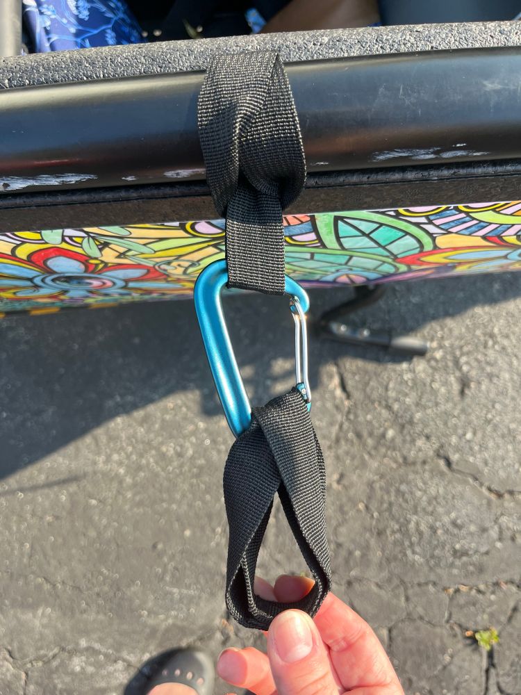 A close-up of a teal carabiner clip attached by a black nylon webbing straps to the frame of an Urban Arrow bucket. There is another strap attached to the bottom in a loop (the hands in the photo are showing the way it loops.)