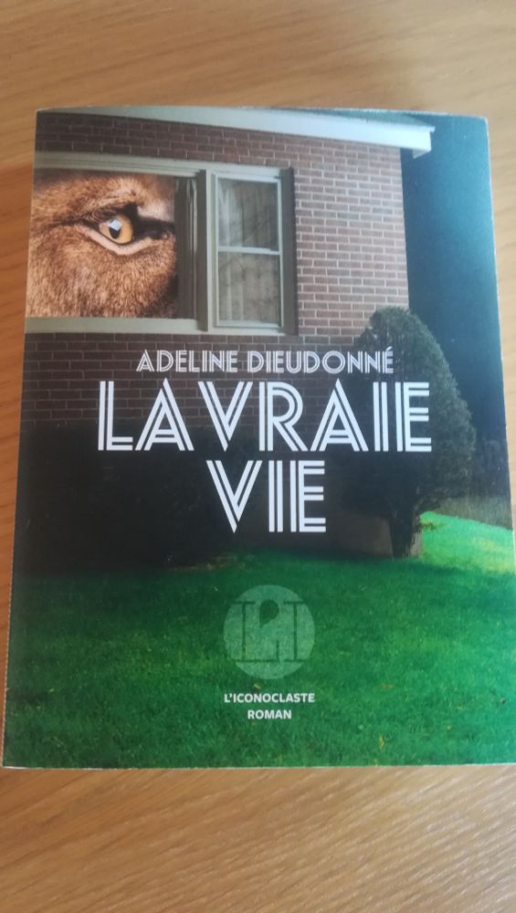 La vraie vie de Adeline Dieudonné. La façade d'une maison, de la pelouse au premier plan. Dans la fenêtre, l'œil d'un félin 