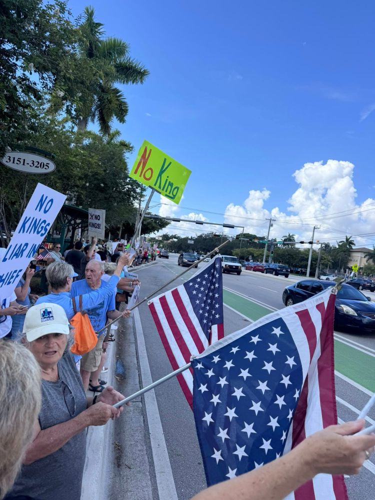 Protesters holding American flags and signs on a city sidewalk saying No Kings. 