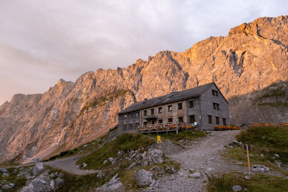 The Lamsenjoch alpine hut standing in front of a large limestone rockwall lit by the early morning sun.