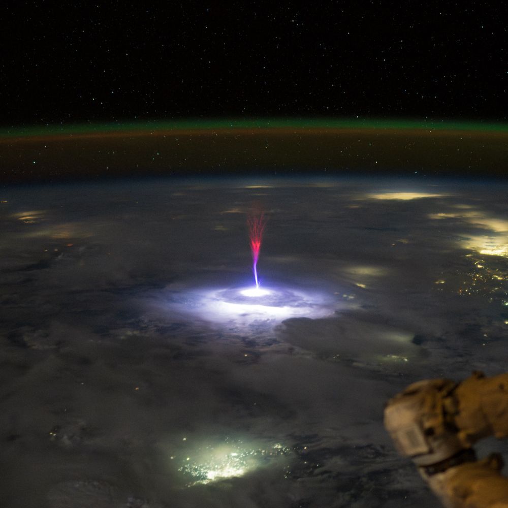 Photograph taken from the International Space Station by astronaut Nichole Ayers, capturing a rare gigantic jet — an upward lightning discharge shooting from a thunderstorm about 90-100 km high into the upper atmosphere. The jet appears as a vivid purple and blue column of light that finely branches out toward the top where it turns into a vivid red color. It's rising from a brightly illuminated cloud top, surrounded by diffuse city lights from Northern Mexico and South Texas. Behind it is the curved limb of Earth with a green band from the airglow layer indicating the edge of the atmosphere. A portion of the space station’s robotic arm is visible in the foreground.
