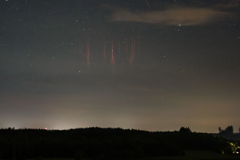 Nighttime image, zoomed in on the sky just above the horizon. Stars are visible above a glowing layer of haze and light pollution. Among the stars are several red sprites, seen as vertical red streaks, some are branching out at the bottom.