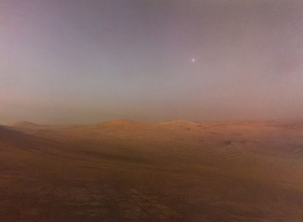 A picture of Mars taken during astronomical twilight (the sun was 13° below the horizon). It shows a dimly lit reddish landscape with some hills at the horizon. Deimos, Mars' smaller moon shines with a small halo in the pale sky above. 