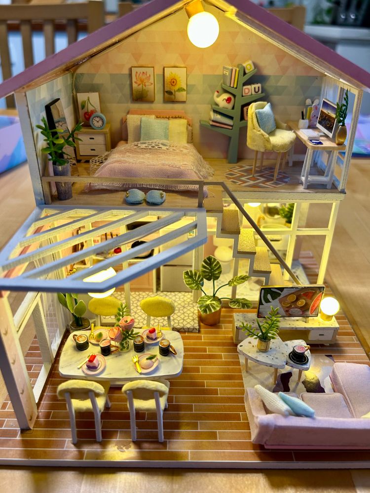 A photo of a tiny two-storey dollhouse (approx. 7x7x7 inches) filled with furniture and homewares