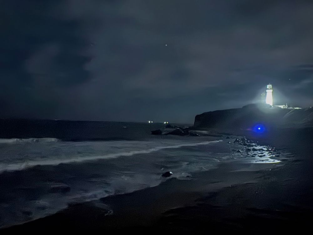 夜の海岸に打ち寄せる波と、岬の上で光を放つ灯台。水平線上には漁船の光と、暗い空にわずかに星が見える。