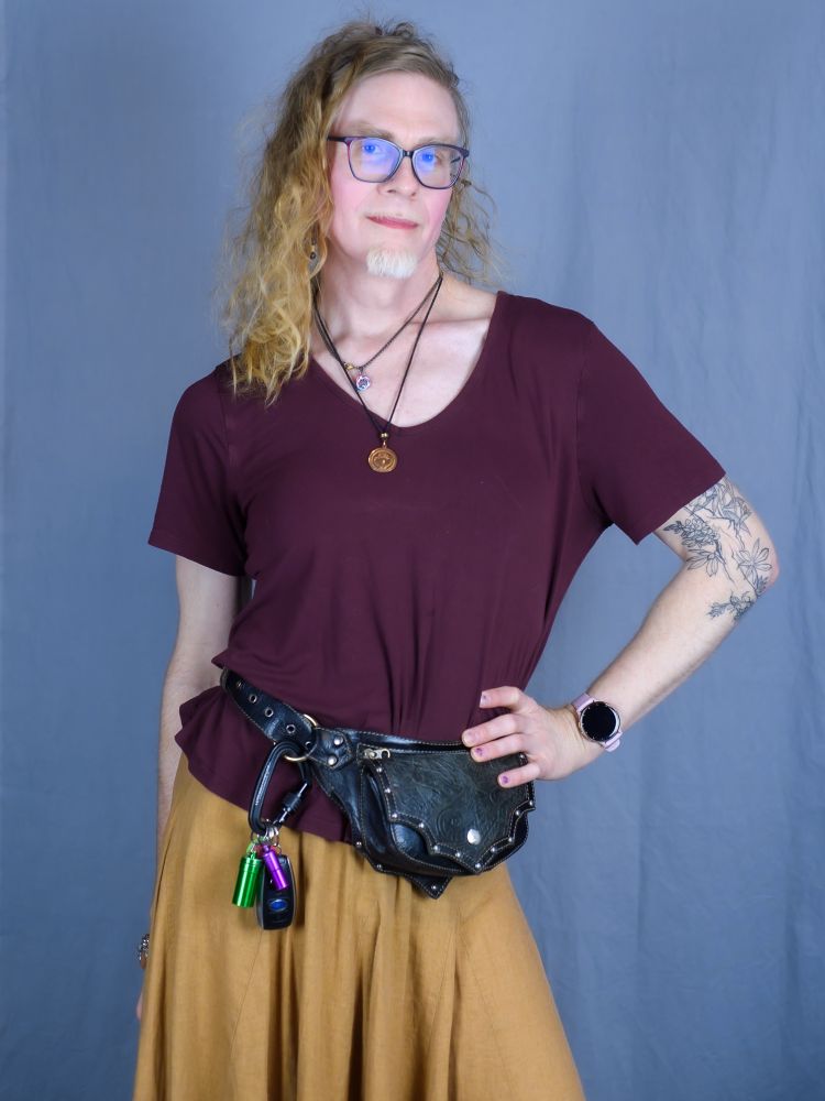 Self portrait of a transfeminine nonbinary person. She has shoulder length red hair and a small white beard. They are wearing a dark red top and a golden brown skirt with a black leather pouch hip belt.