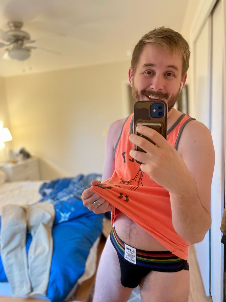 Hyrum smiles while taking a selfie in the mirror and lifting his tank top to display a black and rainbow jock