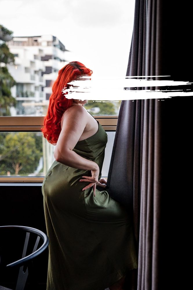 With green fabric cascading down her form, complimented by the vibrant red of her hair, June stands poised in front of a window. Dark curtains frame one side of her, with green forestry appearing behind her. Photography by Jill Kerswill.