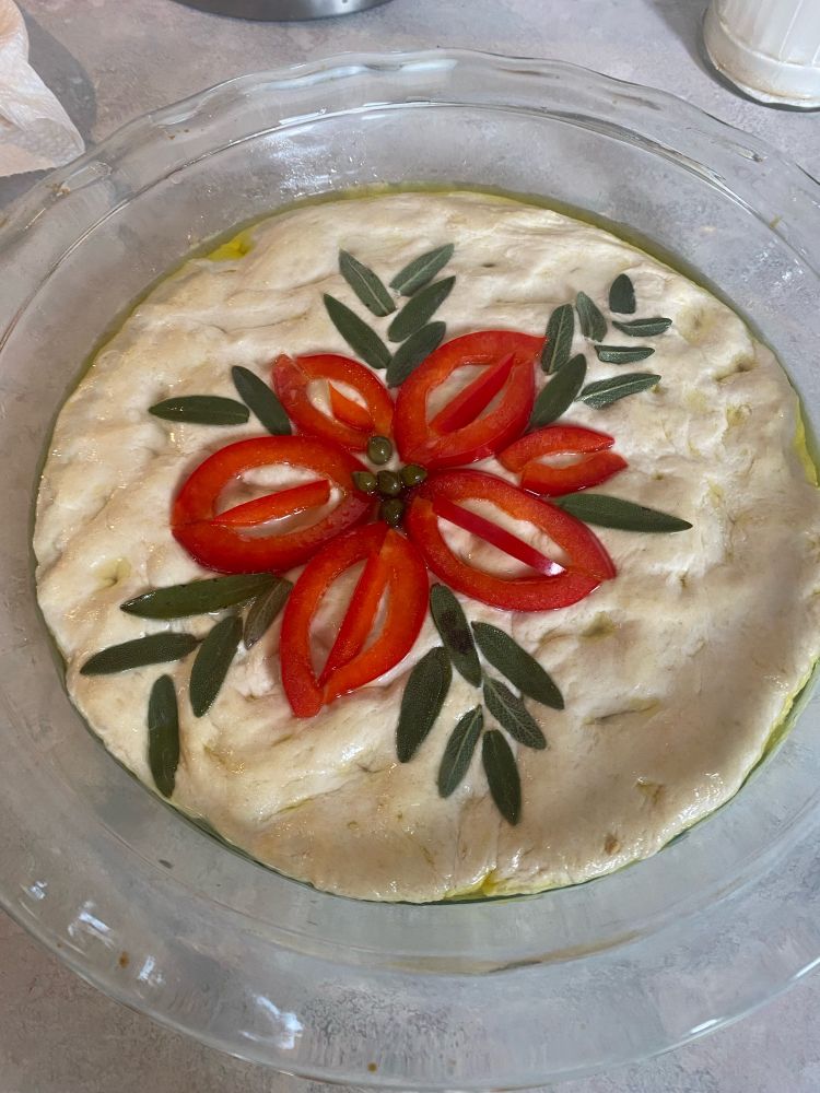 Unbaked focaccia dough with red pepper poinsettia with capers in the middle, surrounded by sage leaves 