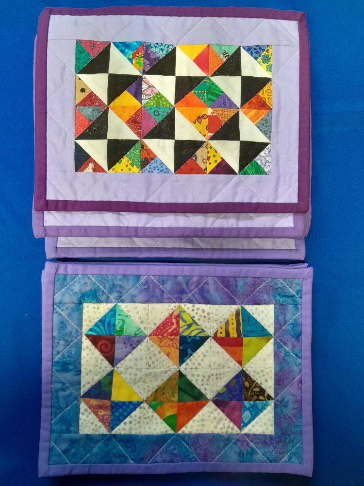 A photo of two stacks of quilted mug rugs. 

On the top, eight mug rugs made of colorful three color quarter square triangles with one side that is either black or white. The borders are lavender, while the bindings are three different shades of purple.

On the bottom, four mug rugs made of batik half square triangles, bordered in a blue-violet batik, bound in light purple.

All of the mug rugs are quilted stitch-in-the-ditch in a diamond pattern.