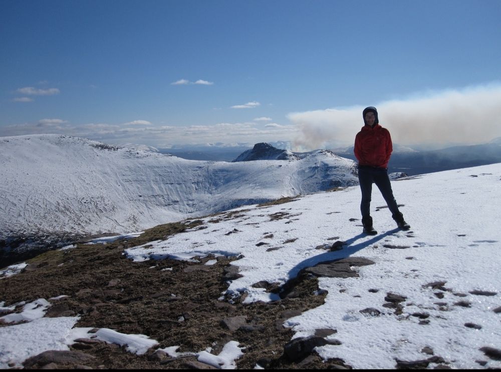 A figure on a snowy slope with smoke from moor fires in the distance 