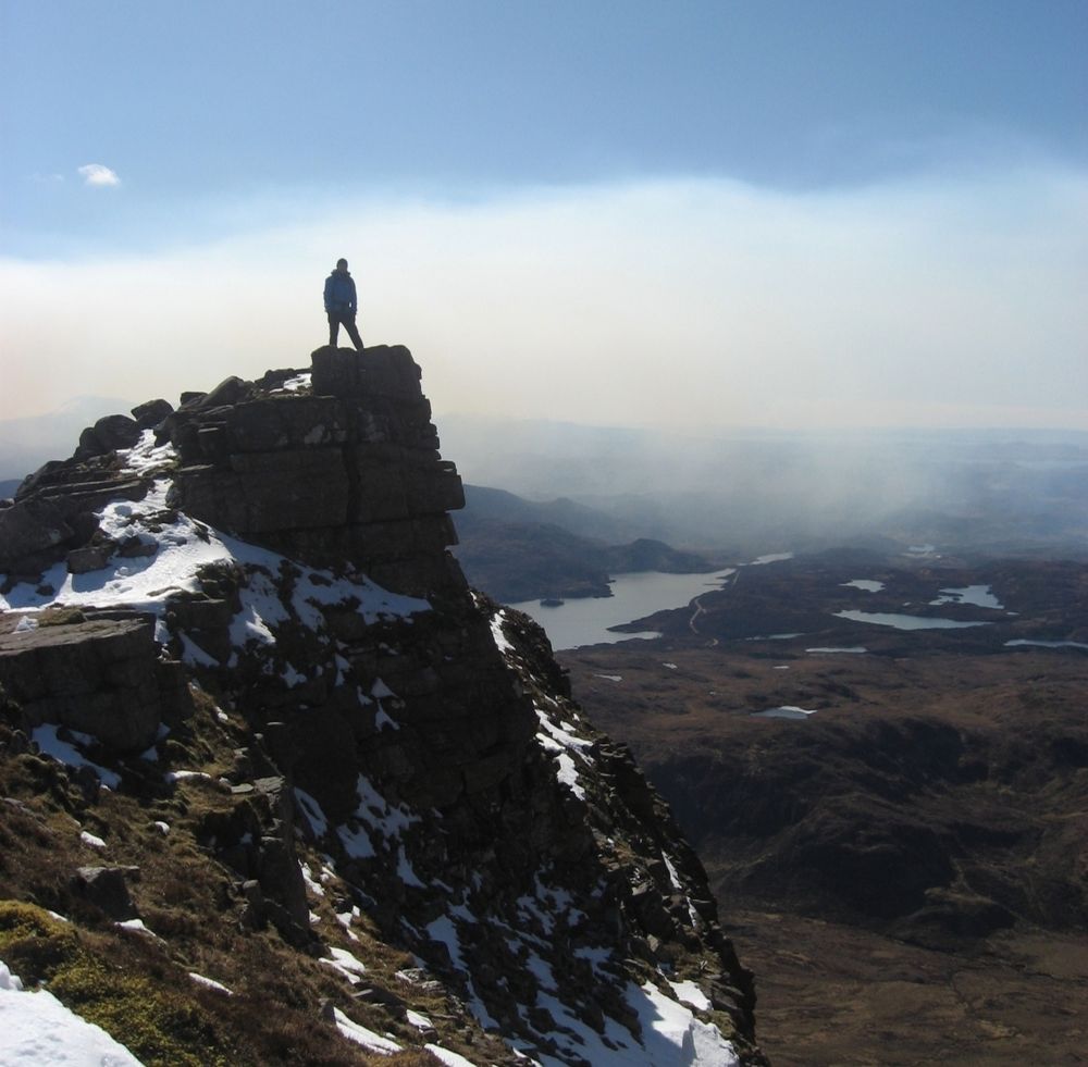 A figure on a rocky peak with a loch behind.