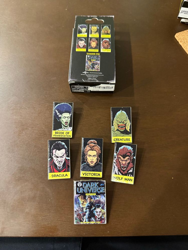 A photo of 6 pins of Universal Monsters on a brown table arranged neatly under a box that the pins originally came in.