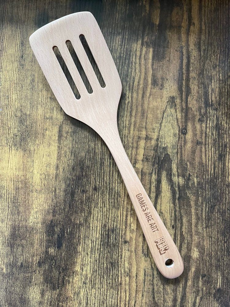 A wooden spatula with the words “games are art” and the Raw Fury logo on the handle. 