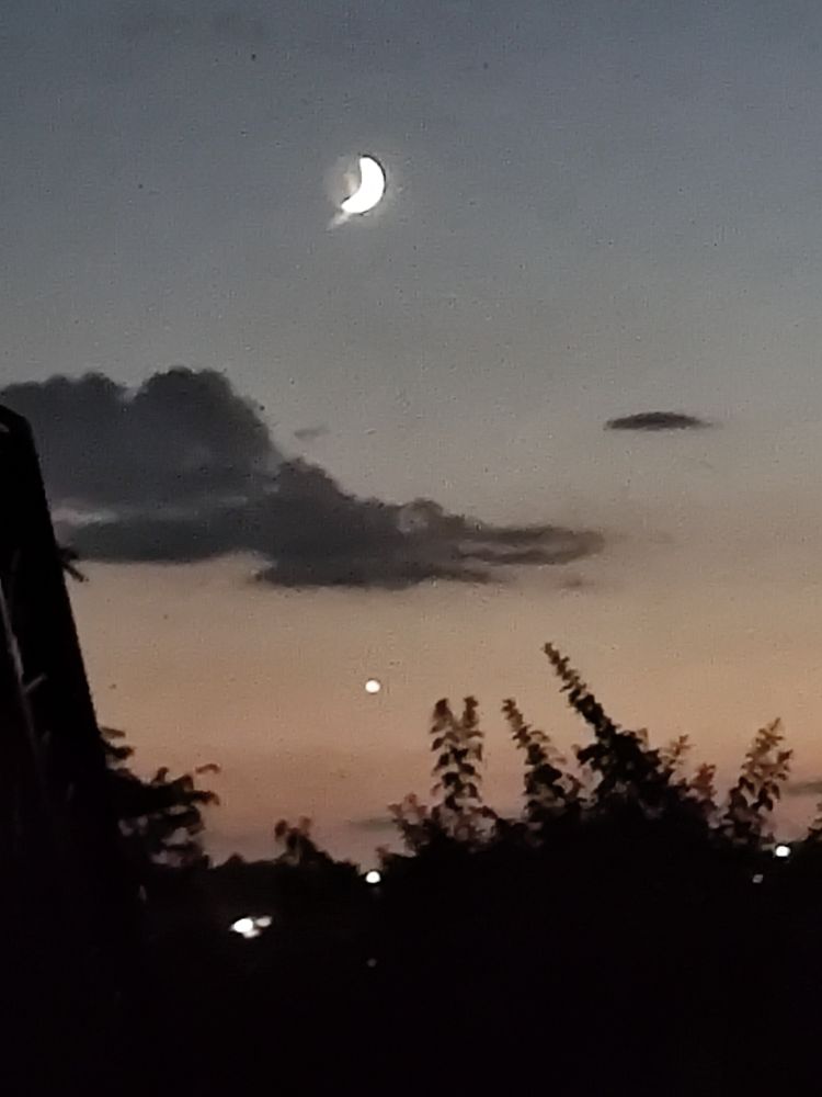 A blurry photo of the sky during nautical twilight after sunset. The (waxing) crescent moon is above a cloud.