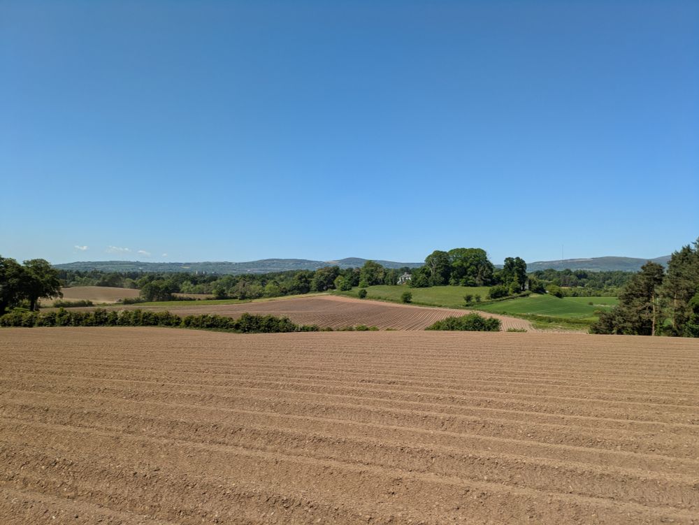 A rolling landscape of ploughed fields fresh sown with potato ridges, drumlins, a historic house in a clump of trees in the distance, set against blue sky on a sunny day.
