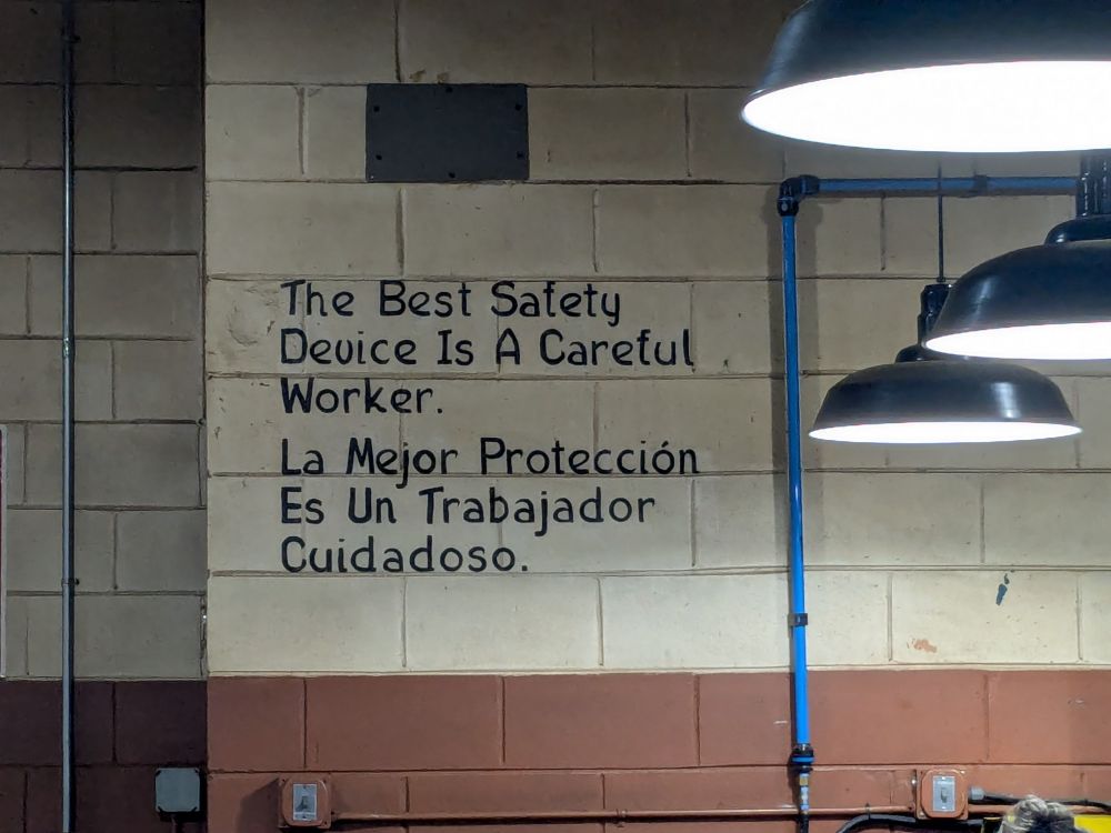 Words hand-painted on a factory wall in an old-fashioned script, in English and Spanish:
The Best Safety Device Is A Careful Worker.
La Mejor Protección Es Un Trabajador Cuidadoso.