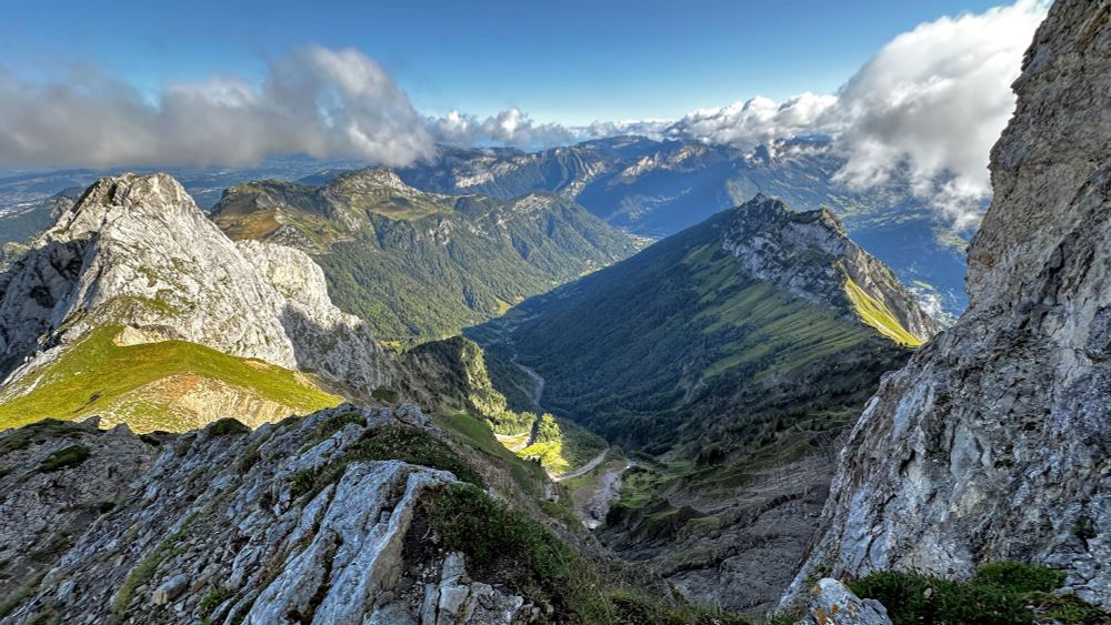 Photo of low mountains marching into the distance, with meadows and forests adorning the slopes. There's a diary farm down there, and higher mountains in the back. The sky is blue, the clouds are fluffy. Frick I wish I was there and not at my computer 