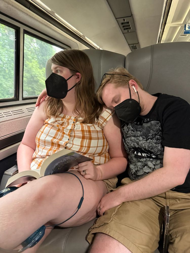 Me, a white trans man with blond hair, sleeping with my head on my wife’s shoulder while they look out the window on the train. We’re both masked.
