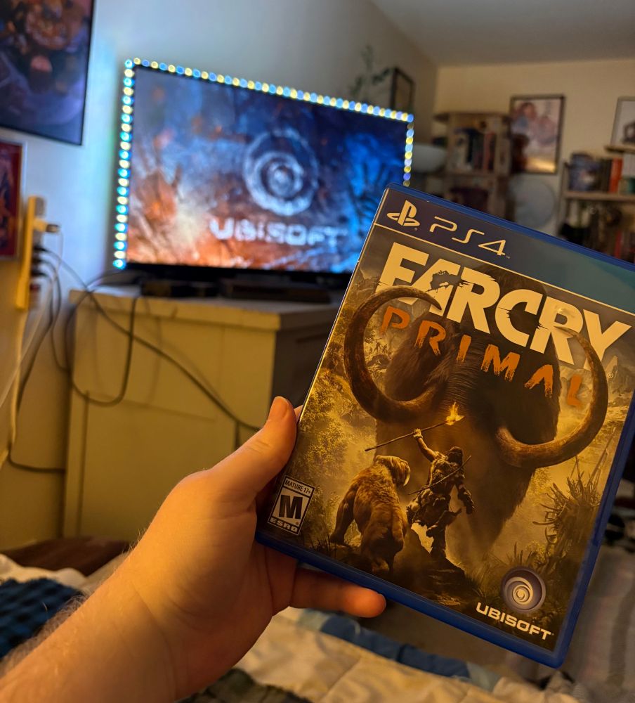 A picture of a white hand holding up a PS4 copy of Farcry Primal, with the Ubisoft logo on the TV screen behind it.
