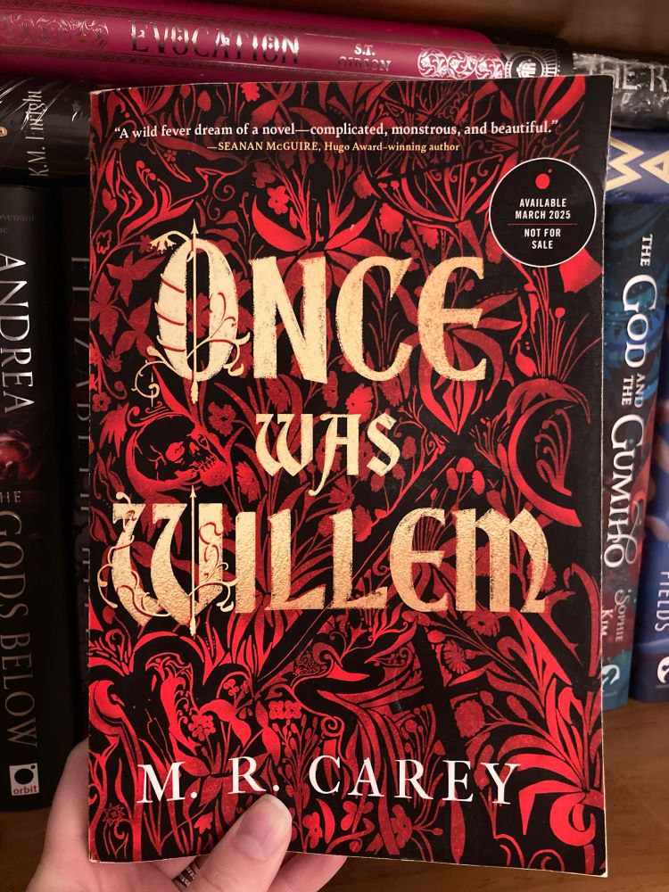 A White hand holding up a book in front of a bookshelf. The book is called “Once Was WIllem” by M.R. Carey. The background is a red and black floral and geometric tapestry-like design.